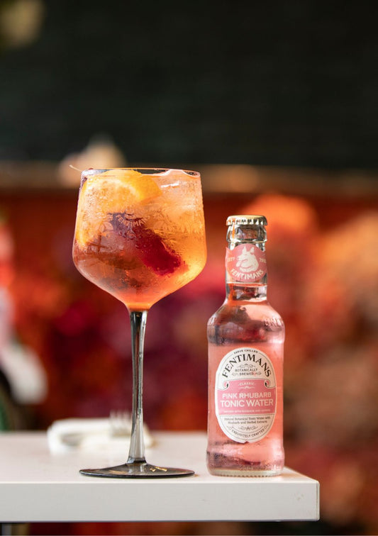 A bottle of Fentimans Rhubarb Tonic Water beside a balloon glass containing the Rhubarb Aperol Fizz cocktail, garnished with orange.