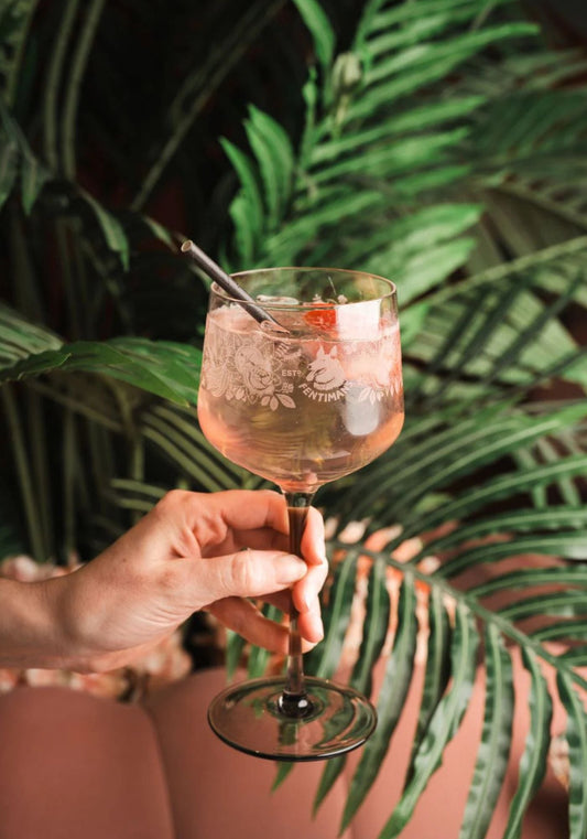 A Fentimans branded balloon glass with pink coloured cocktail garnished with strawberry in someones hand against a palm leaf background