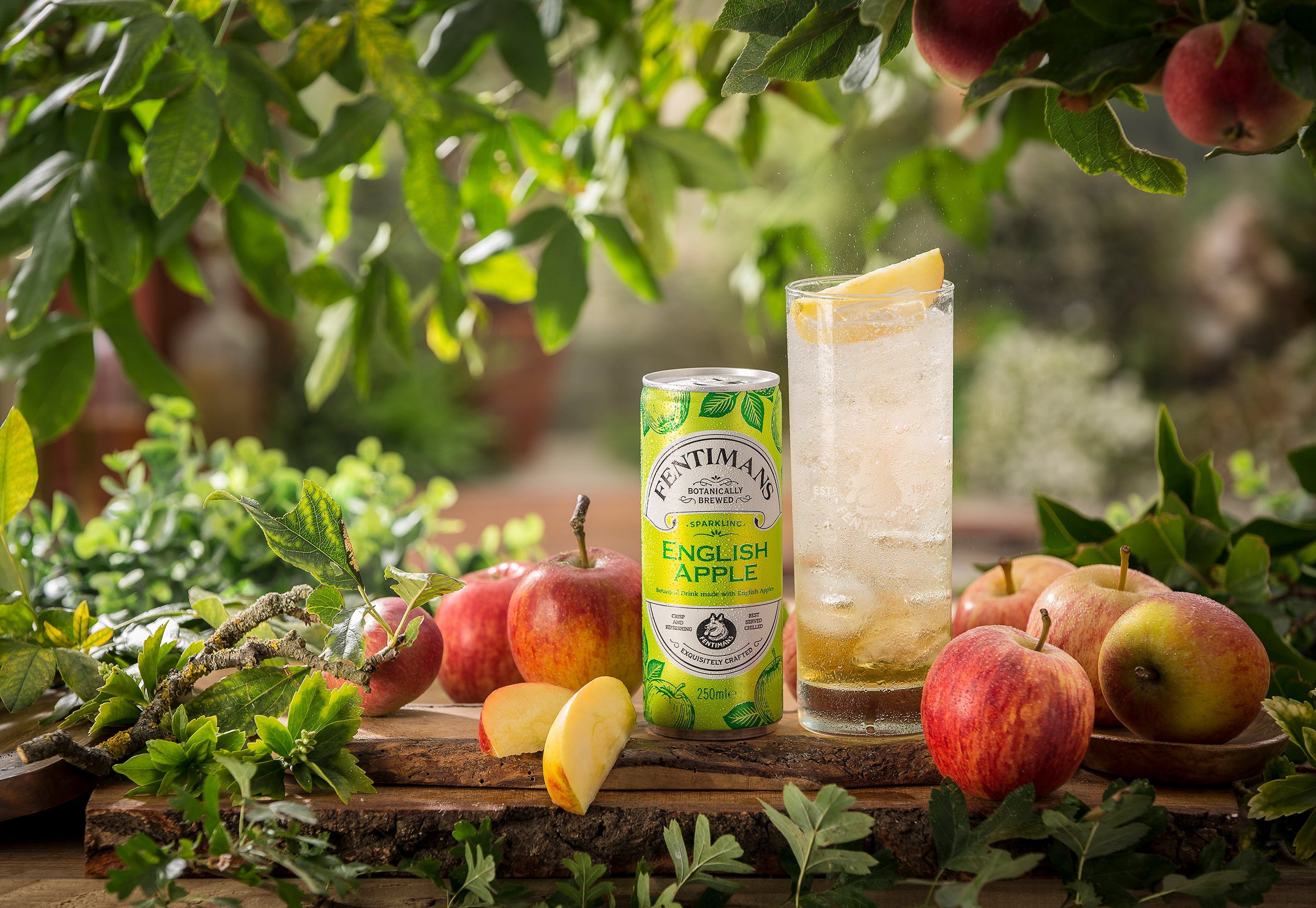 Fentimans English Apple can with a glass filled with Fentimans English Apple surrounded by apples and foliage.
